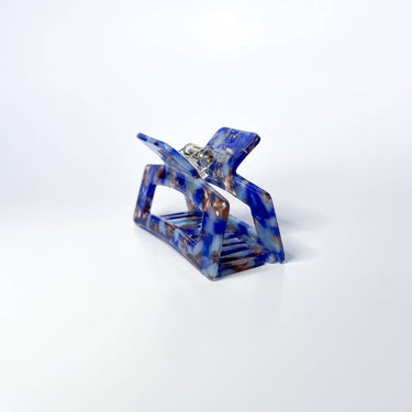 A large, translucent light cobalt blue claw clip is shown against a plain white background. The clip has a rectangular shape with rounded edges and is slightly open, revealing its spring mechanism and gripping teeth.